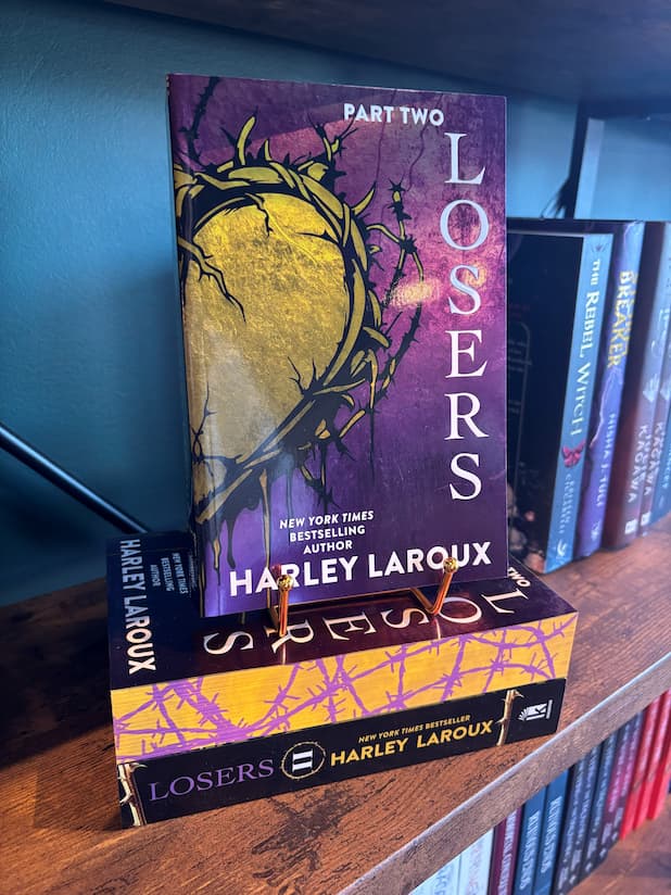 A copy of the book 'Losers: Part Two' by Harley Laroux displayed on a wooden bookshelf, standing upright on a small gold book stand next to other copies of the book.