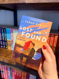 A hand with red nail polish holds a paperback book titled 'Lost and Found' by Tarah DeWitt. The book cover features a blue and peach background of a sunrise with an illustration of a ofa couple embracing in front of a beach with seagulls and grass