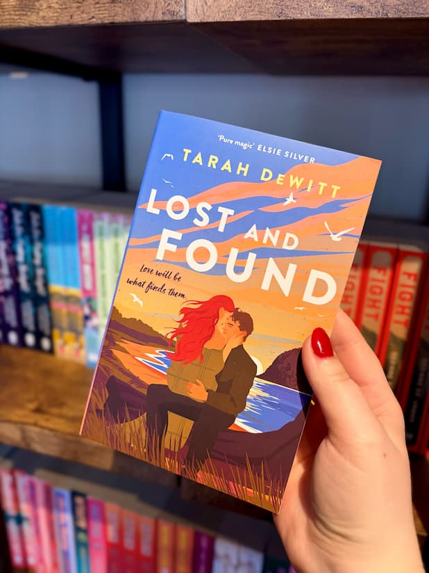 A hand with red nail polish holds a paperback book titled 'Lost and Found' by Tarah DeWitt. The book cover features a blue and peach background of a sunrise with an illustration of a ofa couple embracing in front of a beach with seagulls and grass