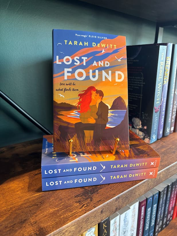 A copy of Lost and Found by Tara Dewitt sits on a gold stand and a stack of the same book on a wooden bookshelf