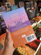A hand with nude nail polish holds the back of a copy of 'Love Means Nothing' by Abra Pressler, featuring a colorful illustrated tennis court scene and the Eiffel Tower in the background with white text.