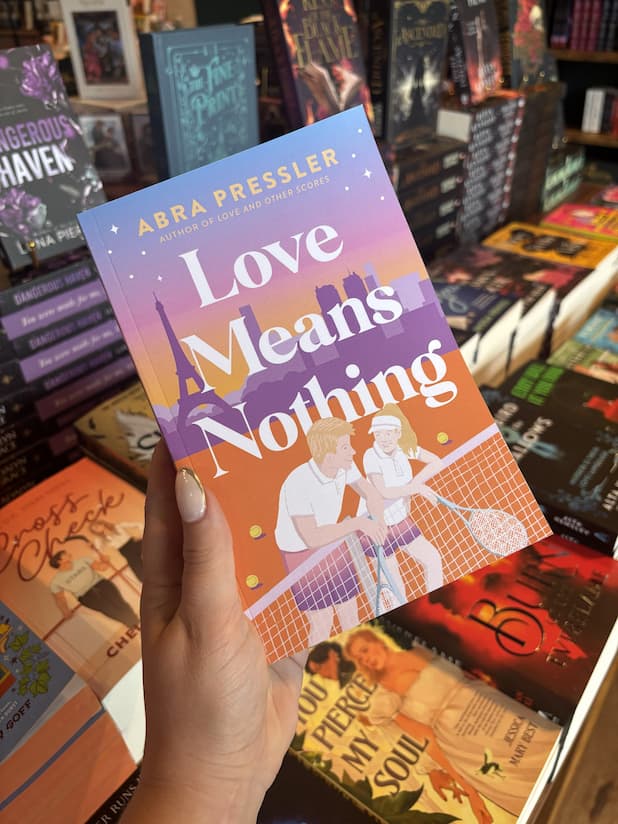 A hand with nude nail polish holds a copy of 'Love Means Nothing' by Abra Pressler, featuring a colorful illustrated tennis court scene with two players, tennis rackets, and the Eiffel Tower in the background.