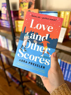 A hand holding a copy of 'Love and Other Scores' by Abra Pressler, featuring two men, one playing tennis, and one wearing an apron and holding a tray of drinks, against a city skyline.