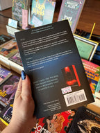 A hand with blue glitter nail polish holds a coy of 'Loving the Wicked' by Rebecca Johnpee, showing the back cover, in a bookstore aisle with other books visible in the background.