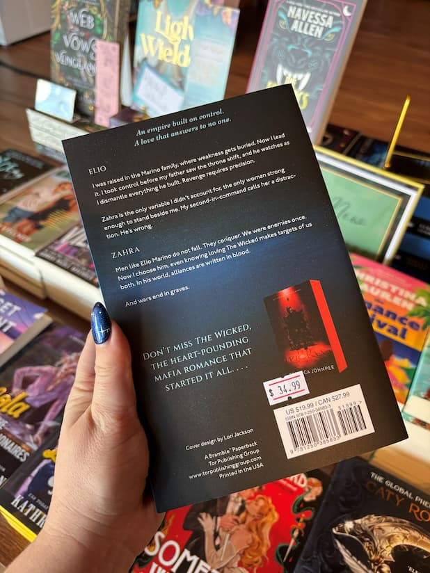A hand with blue glitter nail polish holds a coy of 'Loving the Wicked' by Rebecca Johnpee, showing the back cover, in a bookstore aisle with other books visible in the background.