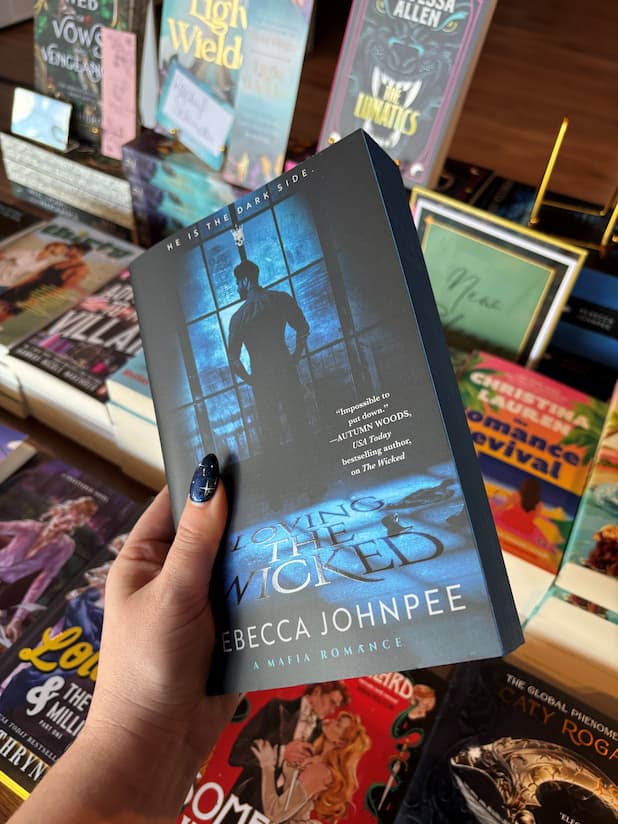 A hand holds the paperback book 'Loving the Wicked' by Becca Johnpee in a bookstore, with other books visible on the shelves in the background.