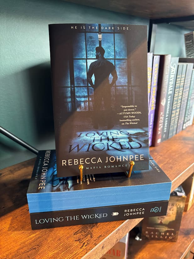 A copy of the book 'Loving the Wicked' by Rebecca Johnfee stands upright on top of another copy of the book on a wooden bookshelf. The cover features a dark silhouette of a man in front of a blue-toned window, with the title in large shadow-looking letters and the author's name in white.