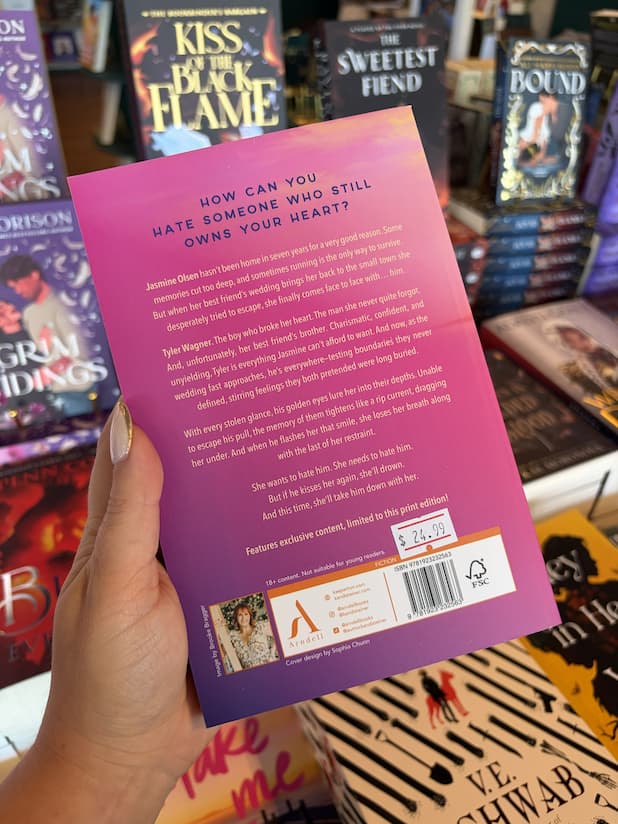 A hand with nude nail polish holding the back of a copy of 'Make Me Hate You' by Kandi Steiner, featuring pink and purple gradient backgroudn with blue and yellow text.