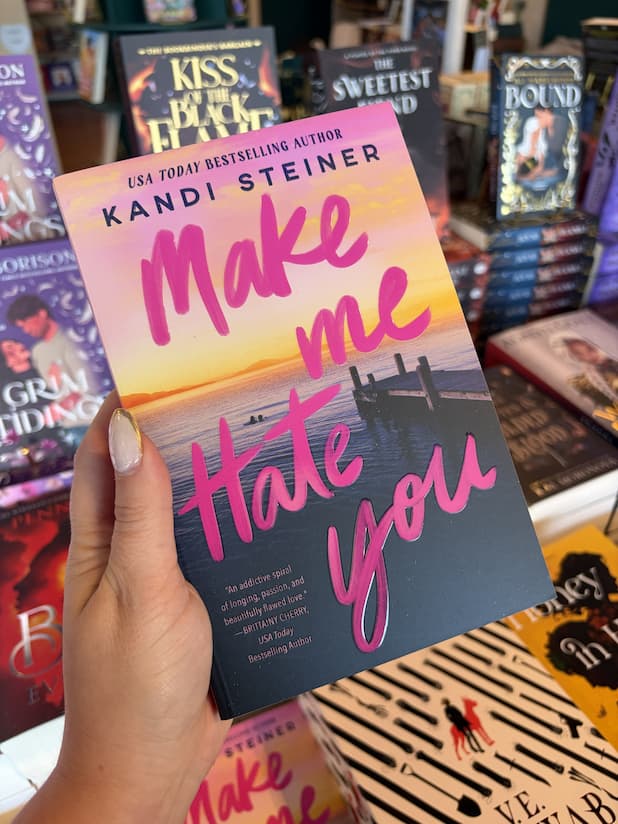 A hand with nude nail polish holding a copy of 'Make Me Hate You' by Kandi Steiner, featuring a pier on a beach at dusk with two people swimming in the water with bright pink text for the title over the image.
