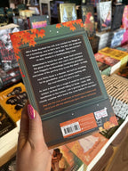 A hand with pink nail polish holds the back of a copy of 'Meet Cute Magic' by Morgan Elizabeth, featuring an illustrated autumn-themed storefront window with white and brown text.