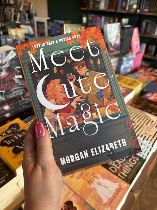 A hand with pink nail polish holds a copy of 'Meet Cute Magic' by Morgan Elizabeth, featuring an illustrated autumn-themed storefront window with a woman and man facing each other holding a heart-shaped potion and a black cat at the bottom.