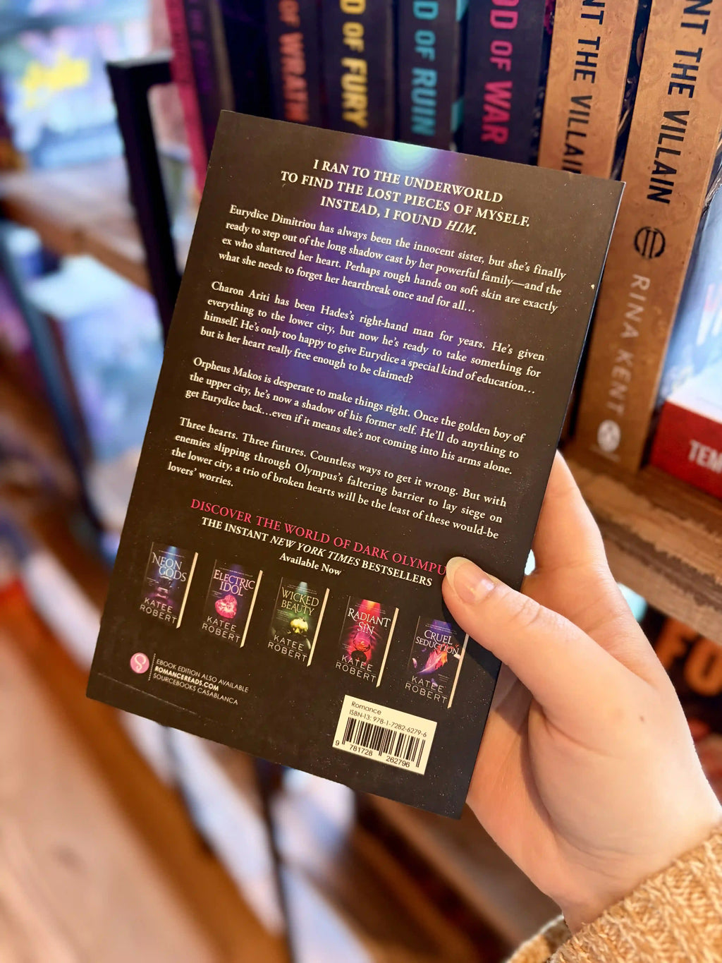 A hand holding the back of a paperback copy of the book 'Midnight Ruin' by Katee Robert. It is a dark and blue cover with orange and white flowers and white text.