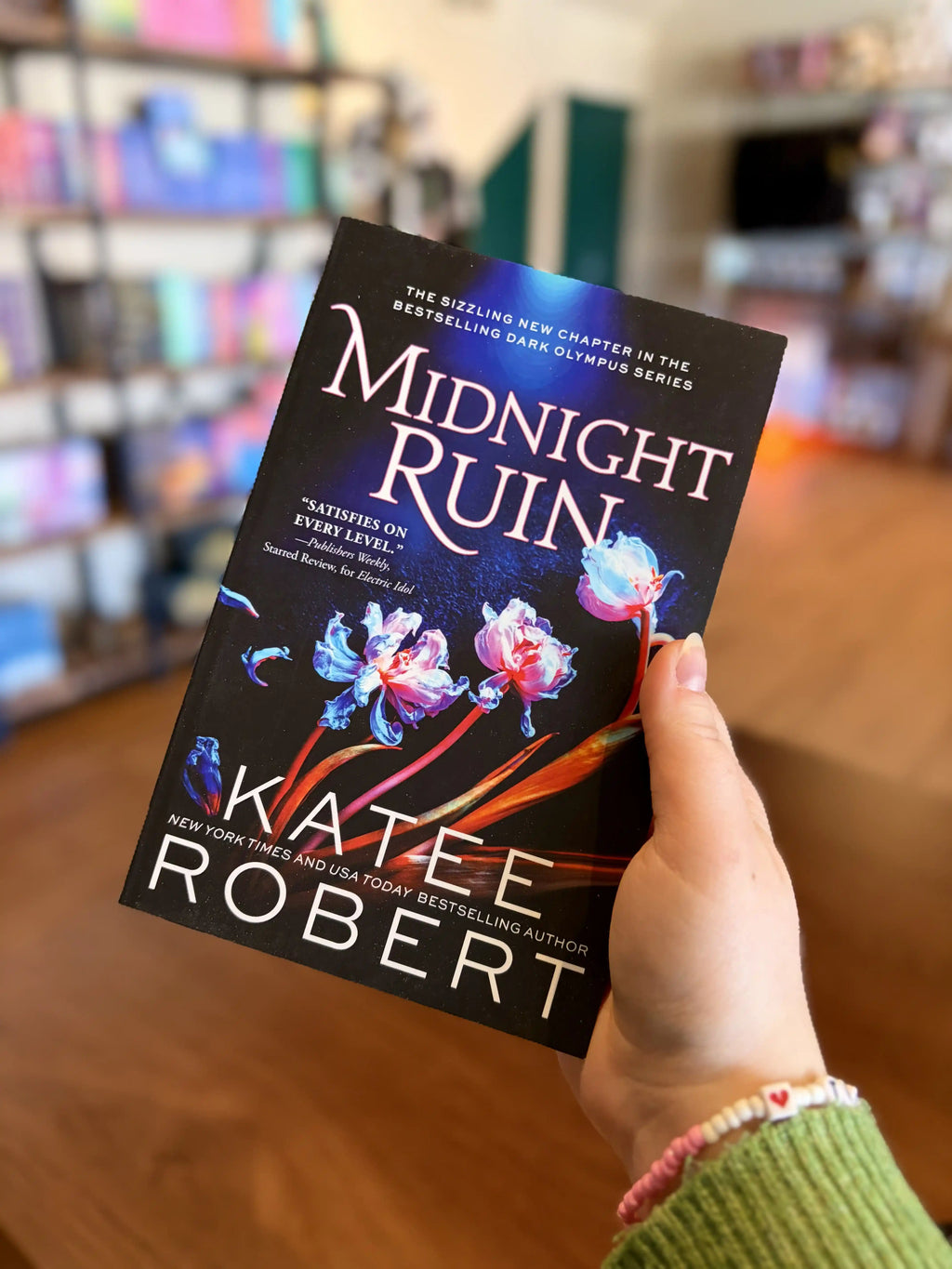 A hand holding a paperback copy of the book 'Midnight Ruin' by Katee Robert. It is a dark and blue cover with orange and white flowers and white text.