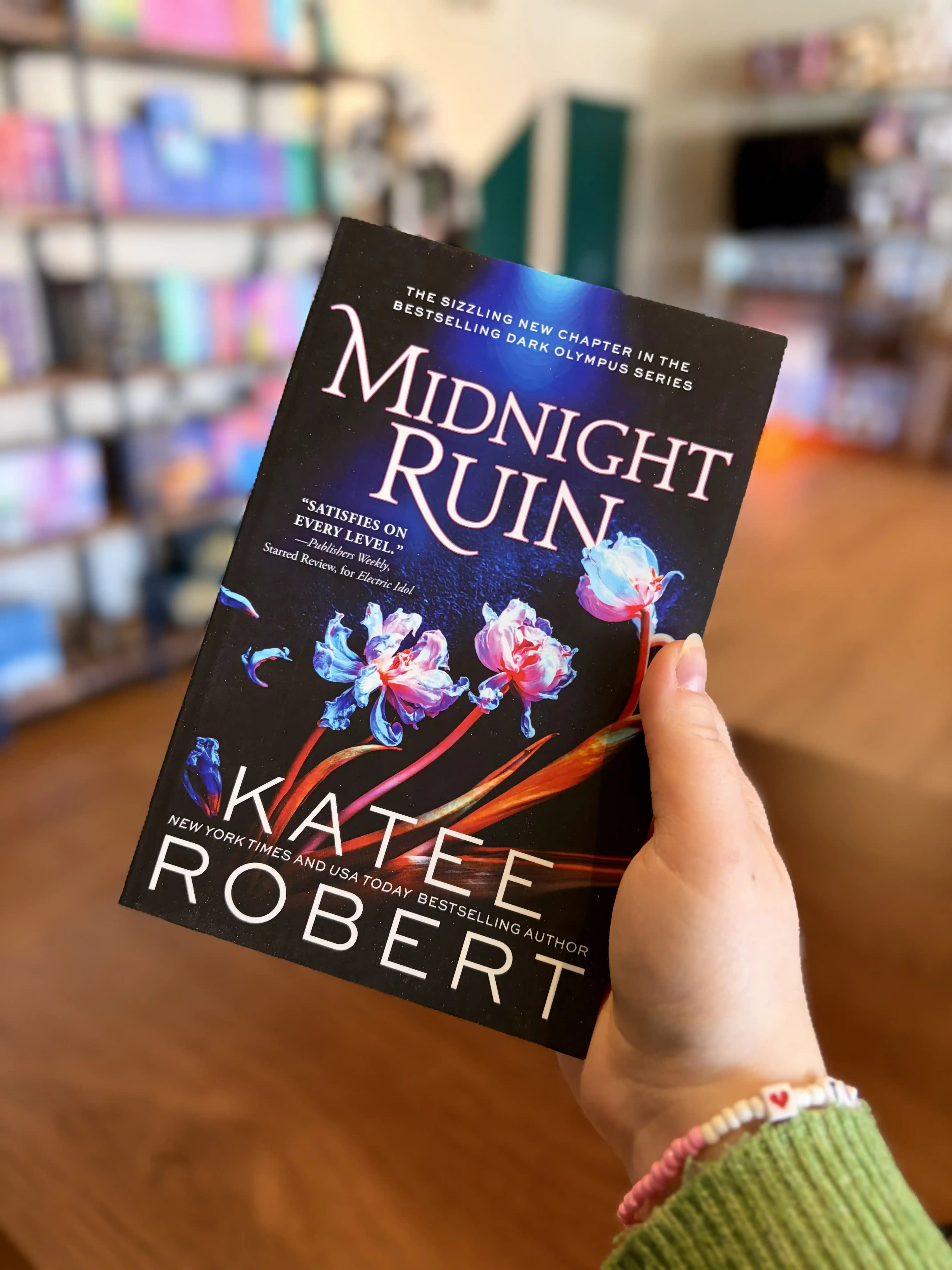 A hand holding a paperback copy of the book 'Midnight Ruin' by Katee Robert. It is a dark and blue cover with orange and white flowers and white text.
