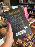 A hand with pink nail polish holding the back of 'Midnight Curfew' by Penelope Douglas, featuring a dark gothic stone archway in the rain with white and yellow text.