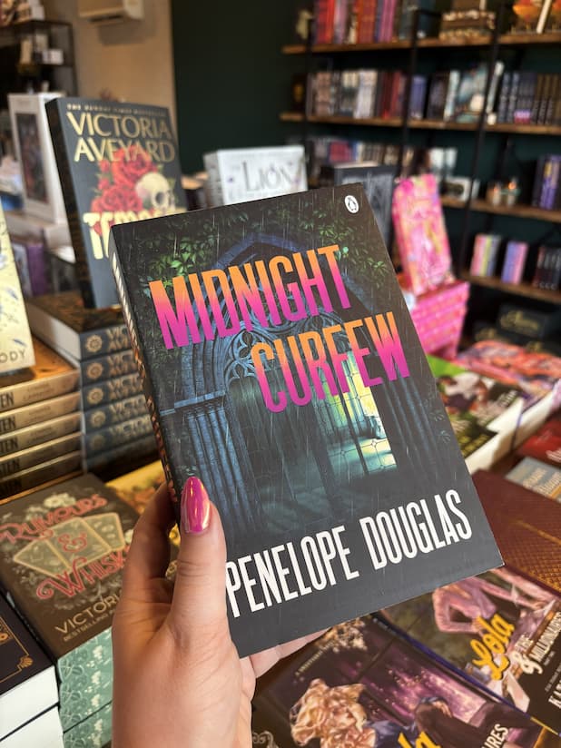 A hand with pink nail polish holds a copy of the book 'Midnight Curfew' by Penelope Douglas in a bookshop. The book cover features a dark gothic illustration with the title in pink and orange gradient text.