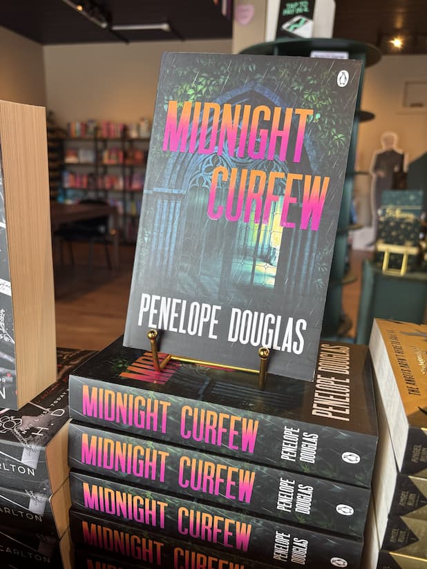 A stack of paperback copies of 'Midnight Curfew' by Penelope Douglas, featuring a dark gothic stone archway in the rain with large gradient title text in pink, orange, and yellow.