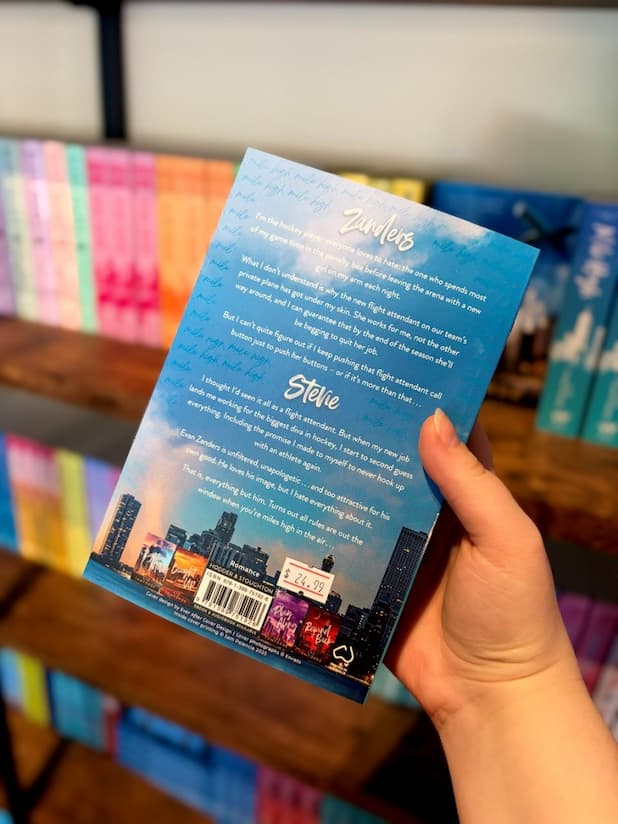 A hand holding the back of a paperback book titled 'Mile High' by Liz Tomforde, featuring a blue sky and city skyline cover with white text, set against a blurred background of bookshelves.
