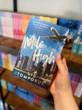 A hand holding a paperback book titled 'Mile High' by Liz Tomforde, featuring a blue sky and city skyline cover with an airplane graphic, set against a blurred background of bookshelves.
