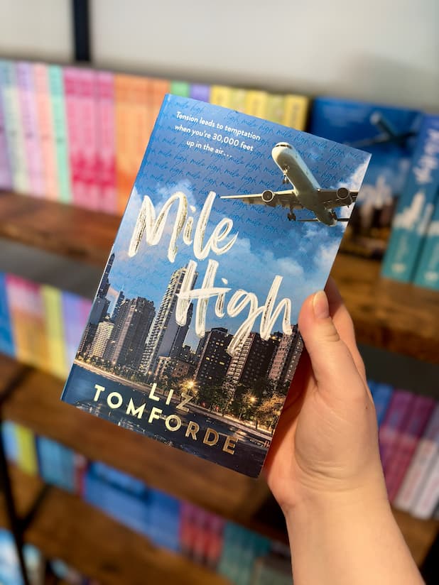 A hand holding a paperback book titled 'Mile High' by Liz Tomforde, featuring a blue sky and city skyline cover with an airplane graphic, set against a blurred background of bookshelves.