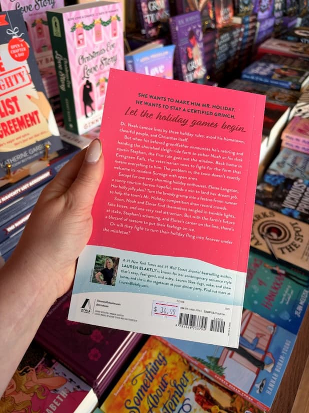A hand with nude nail polish holds the back of a copy of 'Mistletoe All the Way' by Lauren Blakely, featuring a red and white background with white text.