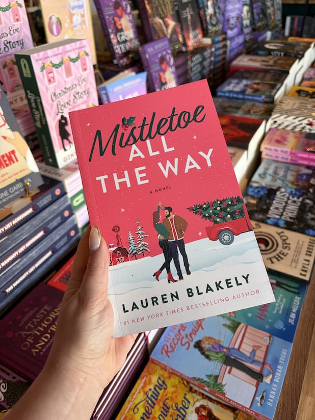 A hand with nude nail polish holds a copy of 'Mistletoe All the Way' by Lauren Blakely, featuring an illustrated couple cuddling under mistletoe in the middle of a snowy small town Christmas scene, with red sprayed edges with holly leaves and berries.