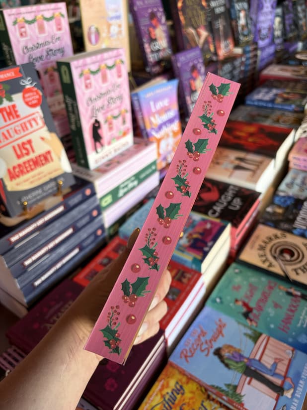 A hand with nude nail polish holds a copy of 'Mistletoe All the Way' by Lauren Blakely, featuring red sprayed edges with holly leaves and berries.
