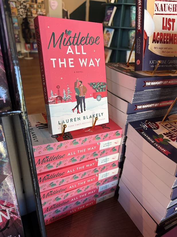 A stack of 'Mistletoe All the Way' by Lauren Blakely, featuring an illustrated couple cuddling under mistletoe in the middle of a snowy small town Christmas scene, with red sprayed edges with holly leaves and berries.