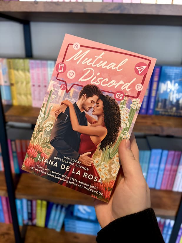 A hand holding a copy of 'Mutual Discord' by Liana de la Rosa, featuring a couple embracing with a cityscape and cacti in the background.