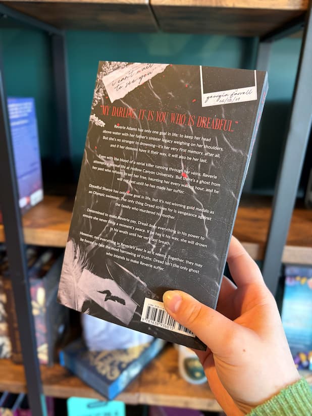 A hand holds the back cover of 'My Dreadful Darling' with white and red font for the blurb on a black background in front of a blurry bookshelf.