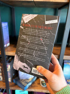 A hand holds the back cover of 'My Dreadful Darling' with white and red font for the blurb on a black background in front of a blurry bookshelf.