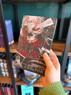 A hand holds a the book cover of 'My Dreadful Darling' with keys, feathers, and text on a dark background with a blurred bookshelf in the background.