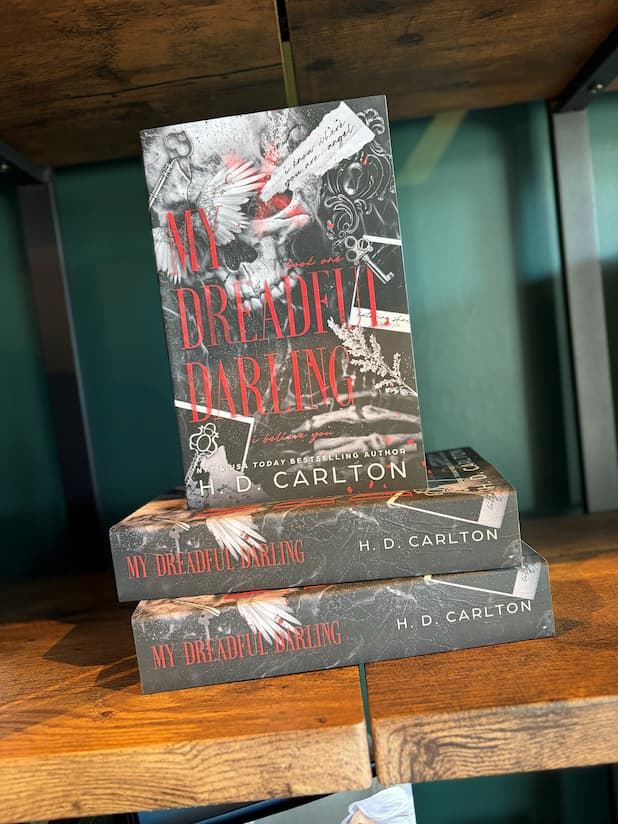 A stack of the book 'My Dreadful Darling' with keys, feathers, and text on a dark background on a wooden bookshelf with a green background.