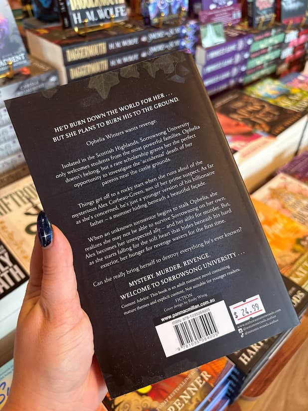 A hand with dark blue nail polish holds a paperback book, showing the back cover of 'Nightshade' by Autumn Woods, in a bookshop with other books blurred in the background.