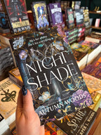 A hand with dark glitter nail polish holds the book 'Night Shade' by Autumn Woods in a bookstore aisle, surrounded by other books on shelves.