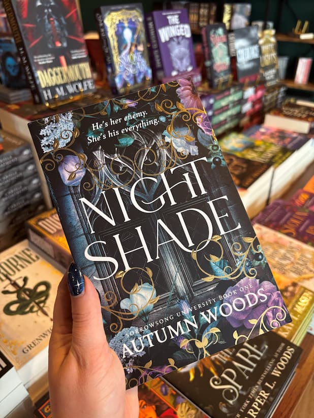 A hand with dark glitter nail polish holds the book 'Night Shade' by Autumn Woods in a bookstore aisle, surrounded by other books on shelves.