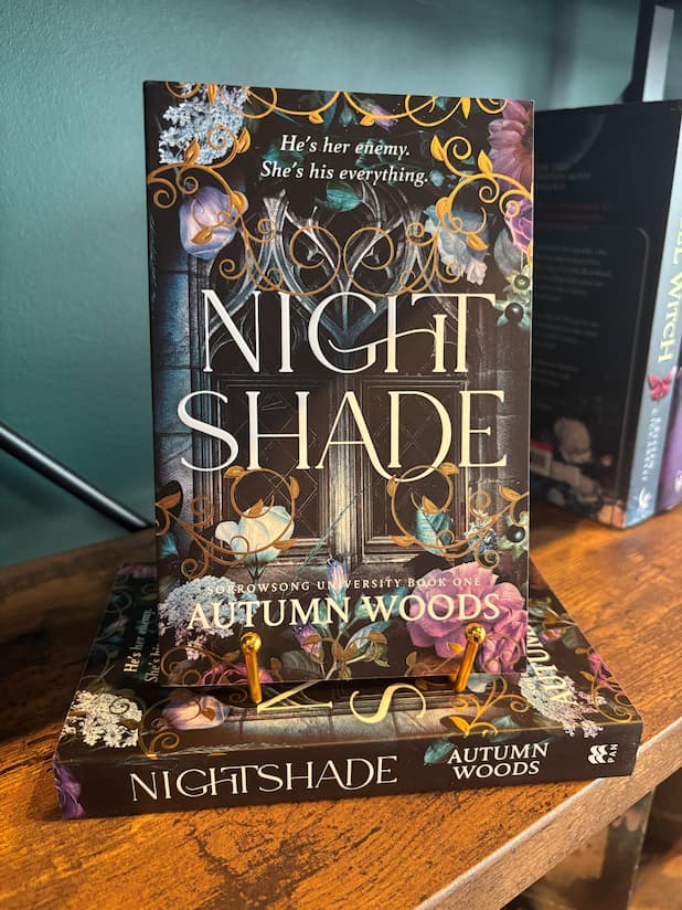 The book 'Nightshade' by Autumn Woods is displayed standing upright on top of its matching book cover, set on a wooden table against a teal wall.