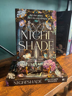 The book 'Nightshade' by Autumn Woods is displayed standing upright on top of its matching book cover, set on a wooden table against a teal wall.