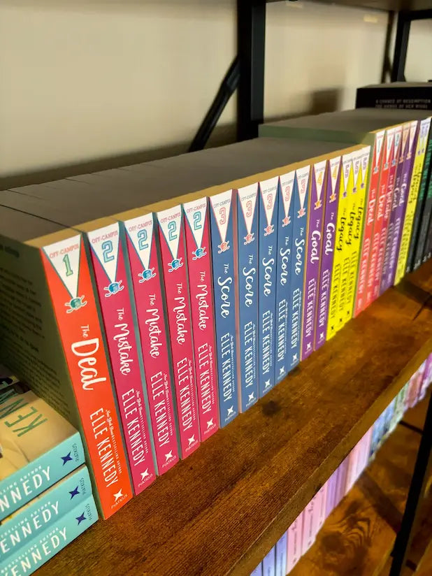 A row of colorful paperback books in a series titled 'Off Campus' by Elle Kennedy displayed on a wooden bookshelf. The spines show different colored chapters: orange, pink, red, blue, purple, yellow, and green.