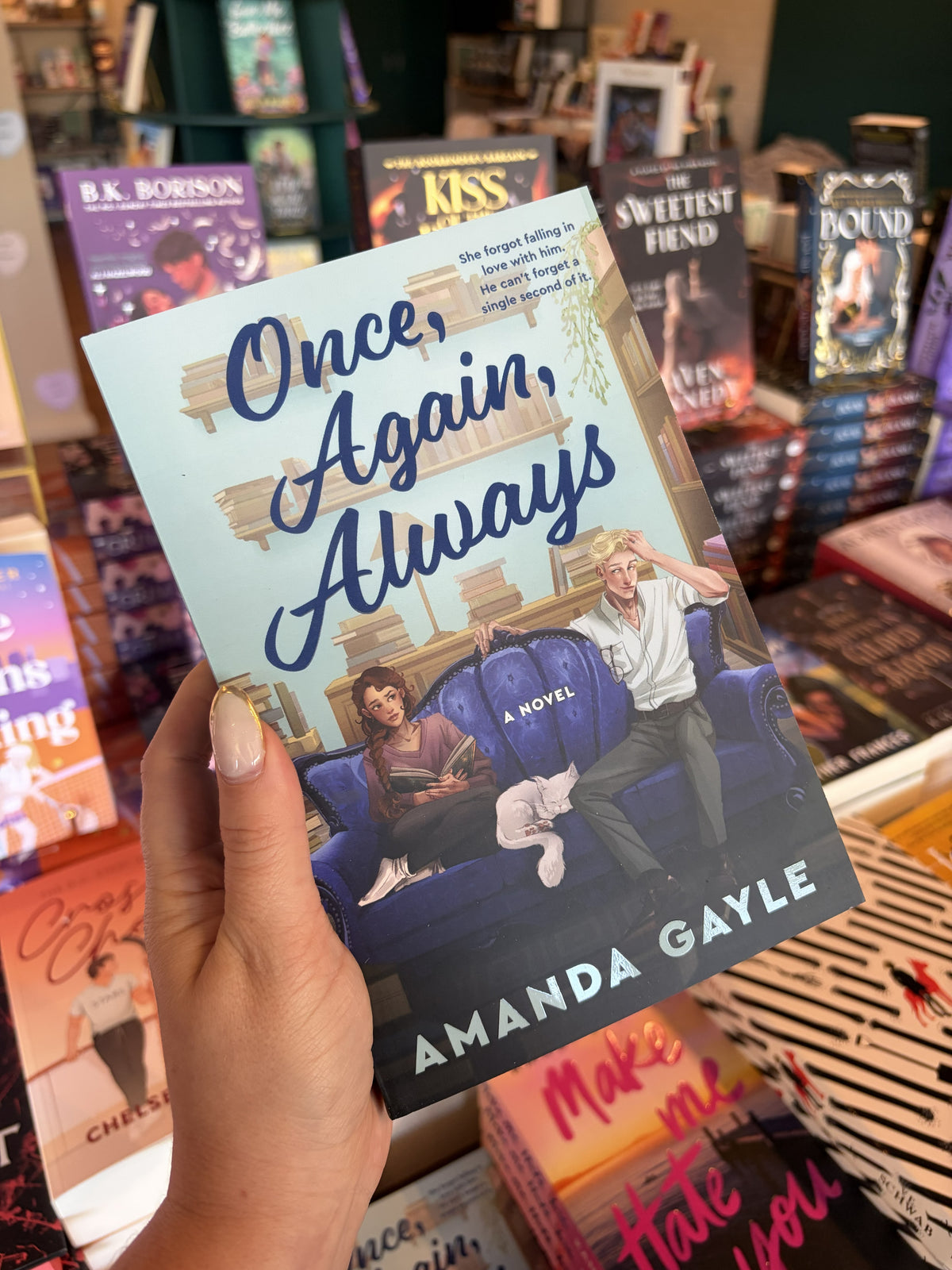 A hand with nude nail polish holding a copy of 'Once, Again, Always' by Amanda Gayle, featuring an illustrated man and woman sitting on opposite ends of a blue sofa with a white cat sleeping between them. They are surrounded by books on shelves and surfaces.