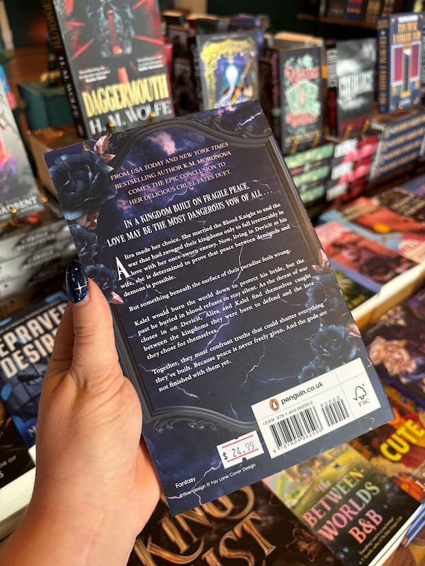 A hand holds the back cover for 'Once Written in Gilded Blood' by K.M. Moronova, Book Two in the Cruel Fates Duet, white text for the blurb is shown against a navy/purple stormy background with a black classical picture frame containing the text. The book is held in front of a table full of other books blurry in the background. 