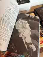 A hand holds open the book 'Once Written in Gilded Blood' by K.M. Moronova, Book Two in the Cruel Fates Duet, an illustration showing the two main characters are shown cuddling in a field.