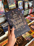 A hand holds the book cover for Once Written in Gilded Blood by K.M. Moronova, Book Two in the Cruel Fates Duet, gilded text overlays blue flowers decorating a classical black and rose gold structure with a sword in the middle. The book is held in front of a table full of other books blurry in the background. 