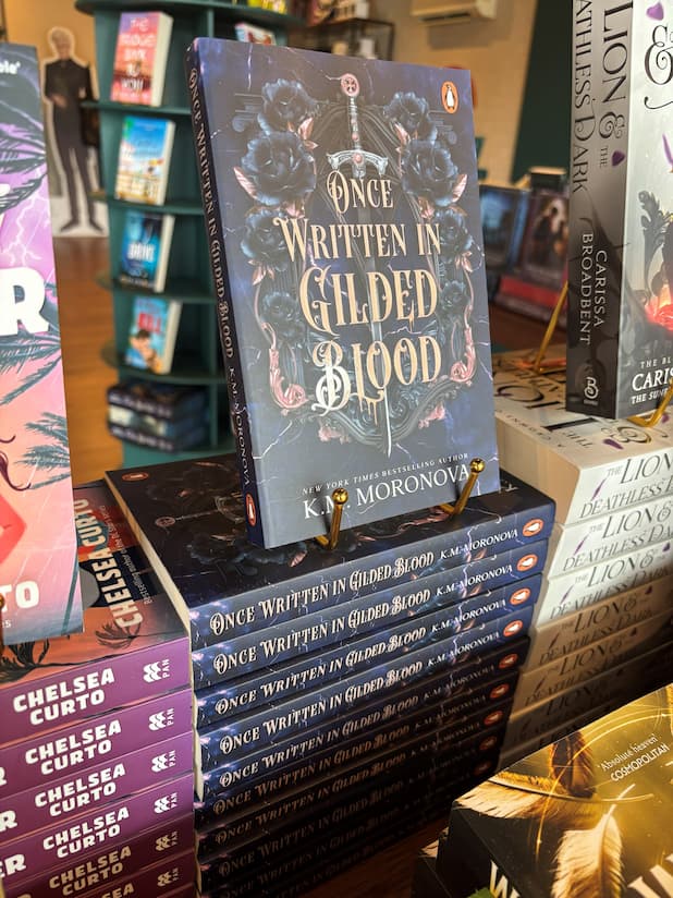 A stack of the book 'Once Written in Gilded Blood' by K.M. Moronova, Book Two in the Cruel Fates Duet, the cover has gilded text overlaying blue flowers decorating a classical black and rose gold structure with a sword in the middle. The stack is located in a bookstore with other books blurry in the background. 