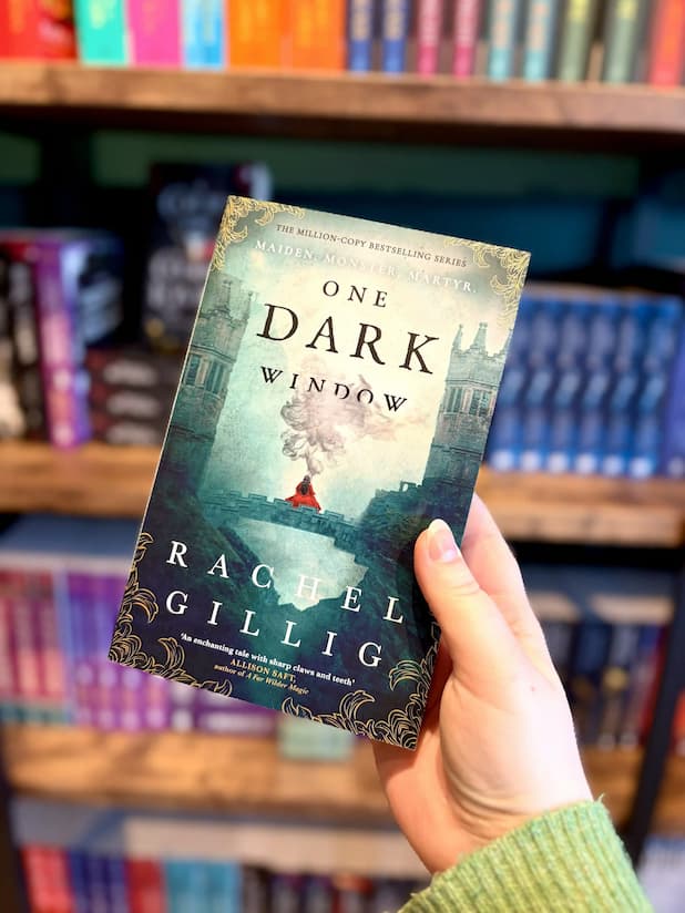 A hand holding a paperback copy of the book 'One Dark Window' by Rachel Gillig, with a blurred bookshelf background. The cover of 'One Dark Window' by Rachel Gillig features a woman in a red dress on a bridge.