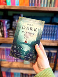 A hand holding a paperback copy of the book 'One Dark Window' by Rachel Gillig, with a blurred bookshelf background. The cover of 'One Dark Window' by Rachel Gillig features a woman in a red dress on a bridge.