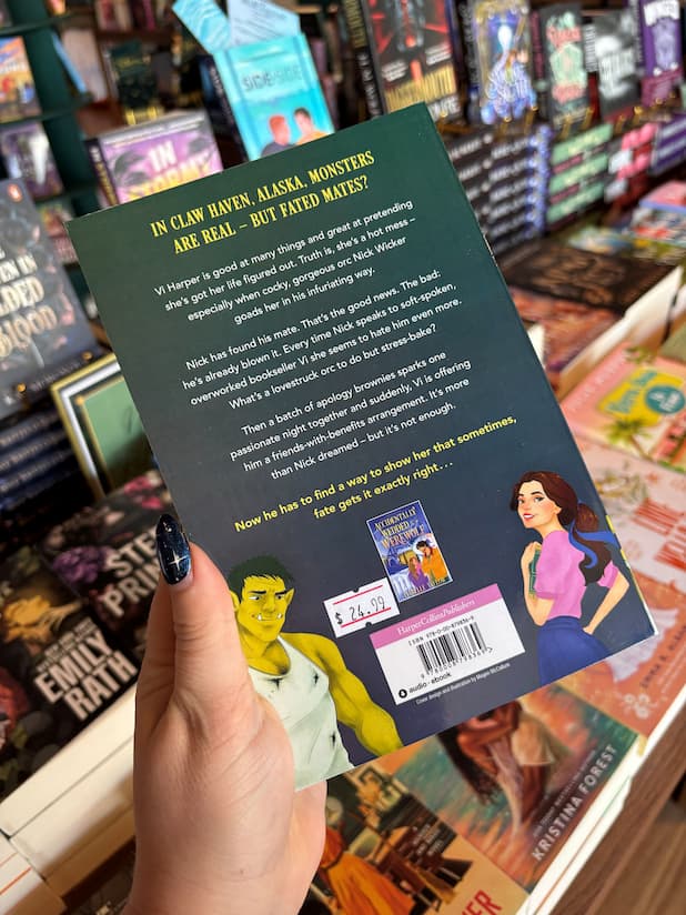 A hand holds the back cover of the book 'Only One for the Orc' by Isabelle Taylor in a bookstore. The dark green cover features illustrations of characters and a barcode, with a blurred background of other books on shelves.