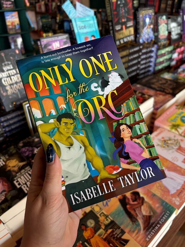 A hand holds the book cover of the book 'Only One for the Orc' by Isabelle Taylor in a bookstore, featuring illustrations of a woman in a pink shirt looking over her shoulder at an orc in a dirty singlet in a library. A white cat with bat wings sits on top of a shelf labelled 'Claw Haven', with a blurred background of other books on shelves.