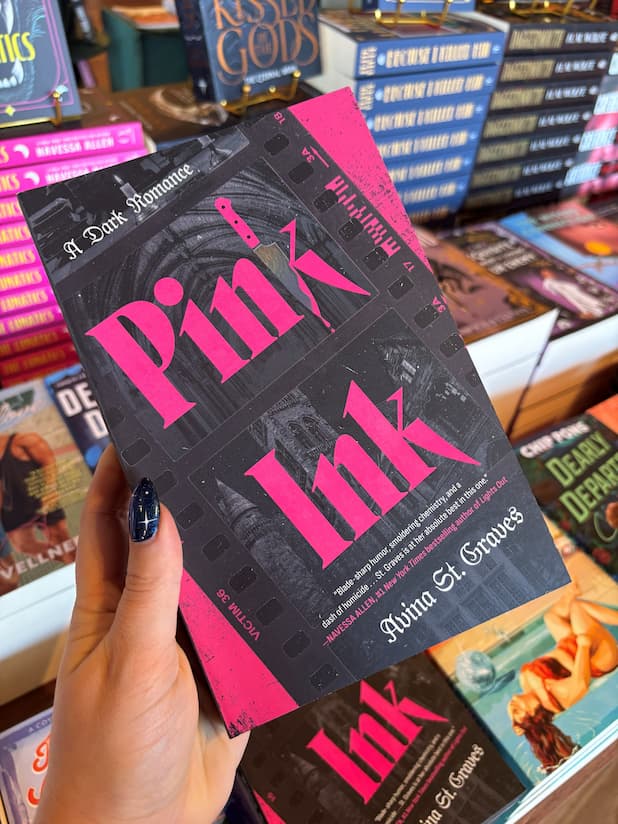 A hand with blue starry nail polish holds a paperback book titled 'Pink Ink' by Avina St. Graves. The book cover features a black university film with pink  text