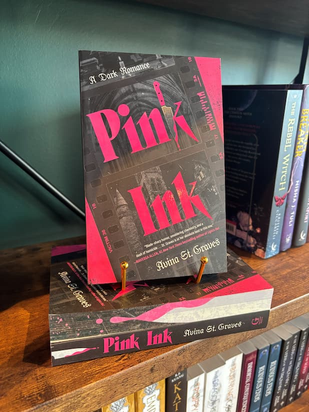 A copy of the book 'Pink Ink' by Avina St. Graves is displayed on a wooden bookshelf, standing upright on a small gold book stand next to other copies of the book.
